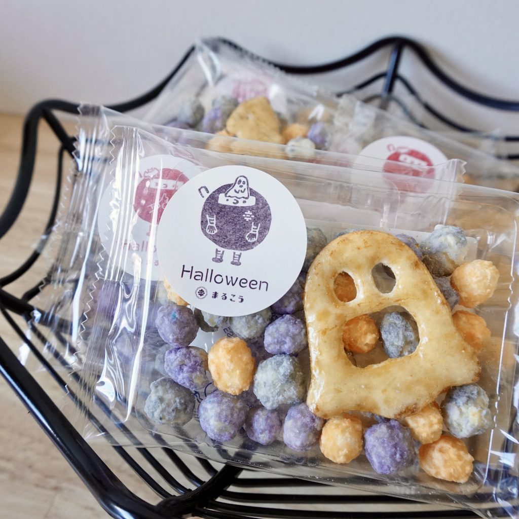 「Halloween まるこう」のシールが貼られた透明袋に入った、おばけの形をした醤油味あられと、黒・黄・紫色の丸い砂糖がけあられ